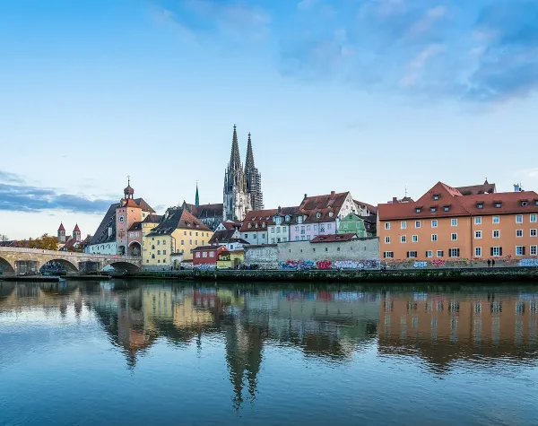 Altbauhäuser, Brücke und Dom spiegeln sich im Wasser in Regensburg unter blauem Himmel.