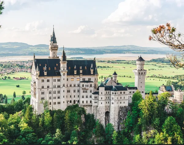 Schloss Neuschwanstein in grüner Landschaft des Ostallgäus, umgeben von Bäumen und Hügelpanorama.