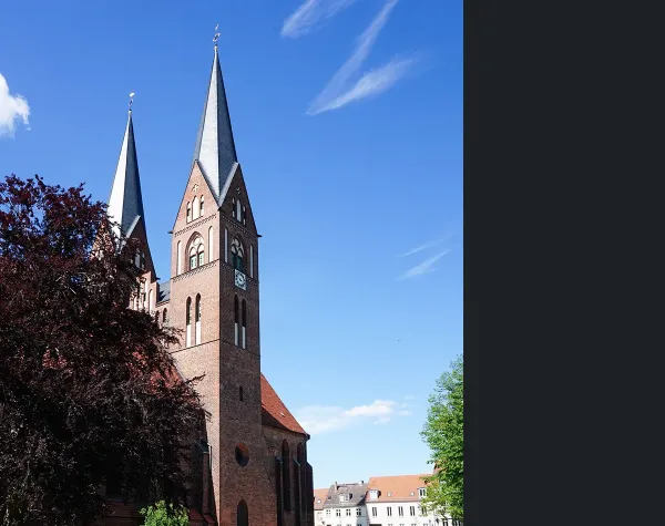 Backsteinkirche in Ostprignitz-Ruppin mit zwei Türmen bei blauem Himmel, umgeben von Bäumen und Häusern.