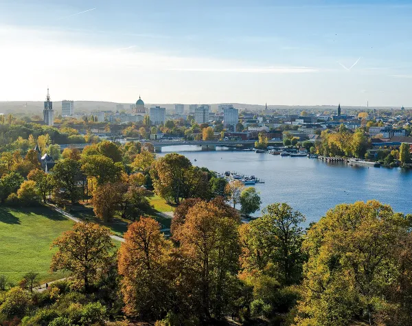 Landschaft in Potsdam-Mittelmark mit Fluss, Bäumen und Gebäuden im Hintergrund bei klarem Himmel.