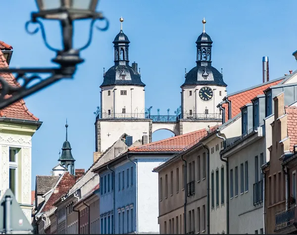 Stadtansicht mit historischem Gebäude und Türmen im Hintergrund, blauer Himmel darüber.