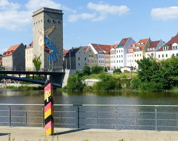 Brücke über Fluss, bunte Häuserreihe, Turm mit großem Wandgemälde, grenznahes Gebiet, sonniger Himmel.