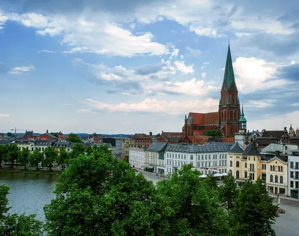 Stadtpanorama von Schwerin mit Dom und umliegenden historischen Gebäuden am Wasser, grünes Laub im Vordergrund.