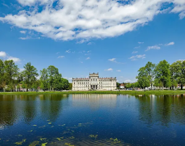 Schloss Ludwigslust an einem sonnigen Tag, reflektiert in einem Teich, umgeben von grünen Bäumen und klarem blauen Himmel.