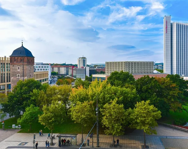 Städtische Skyline von Chemnitz mit Bäumen im Vordergrund, historischen und modernen Gebäuden im Hintergrund.