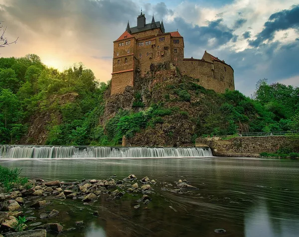 Burg Kriebstein auf einem Felsen über einem Fluss im Sonnenuntergang, umgeben von bewaldeter Landschaft in Mittelsachsen.
