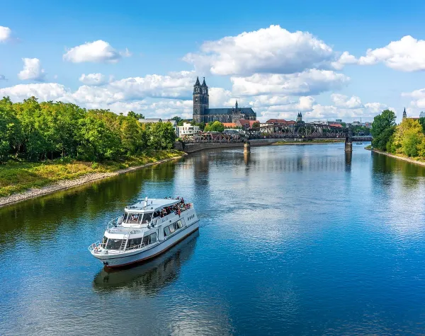 Weiße Ausflugsbootfahrt auf der Elbe, im Hintergrund der Dom von Magdeburg und eine historische Brücke unter blauem Himmel.