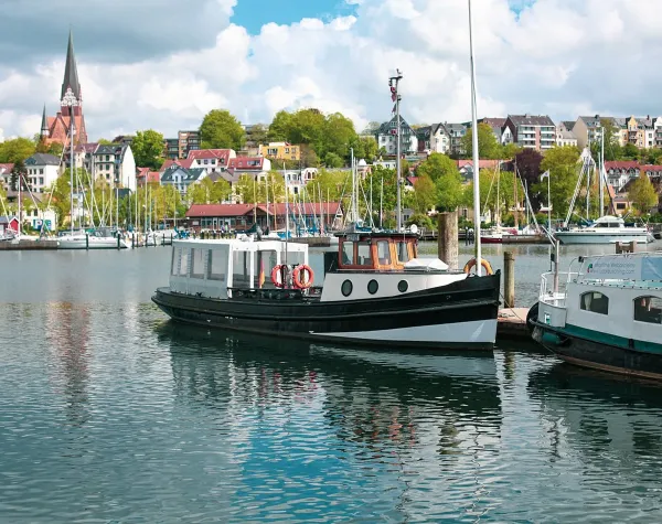 Schiffe im Hafen von Flensburg, im Hintergrund bunte Gebäude und Kirche vor bewaldetem Hügel.