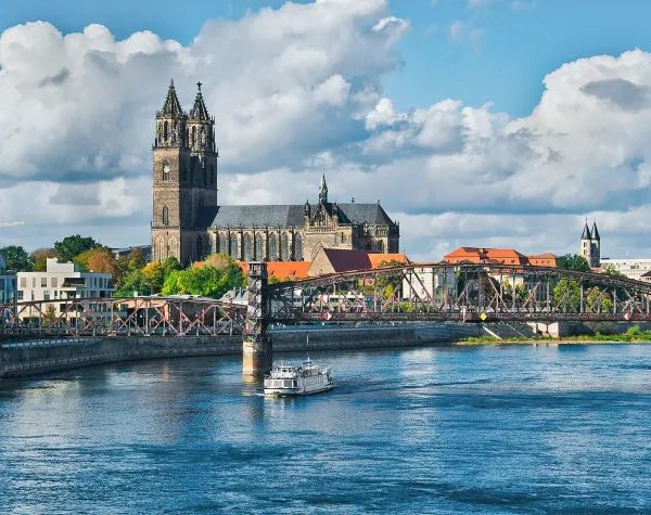 Historische Kirche in Magdeburg, Deutschland, hinter einer Brücke am Flussufer, umgeben von Stadtarchitektur.