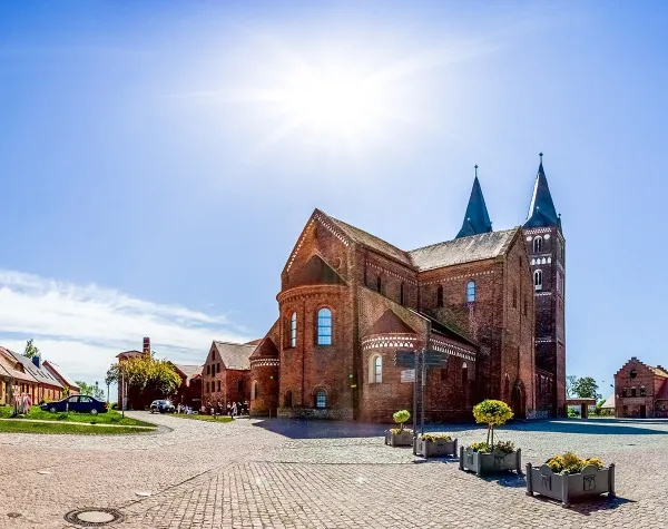 KBackstein-Kirche mit Doppeltürmen in Jerichow unter sonnigem Himmel, umgeben von Kopfsteinpflaster und Pflanzen.
