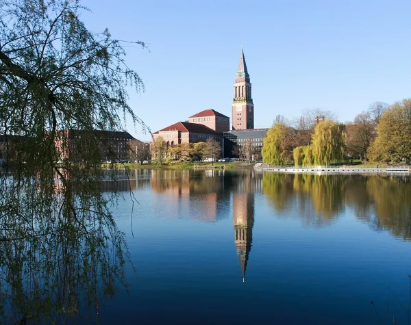 Kirchturm und Gebäude spiegeln sich im See, umgeben von Bäumen in Kiel.