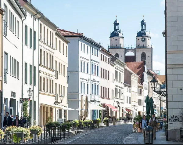 Straße in Wittenberg mit historischen Gebäuden und der Stadtkirche im Hintergrund, Menschen spazieren entlang der Geschäfte.
