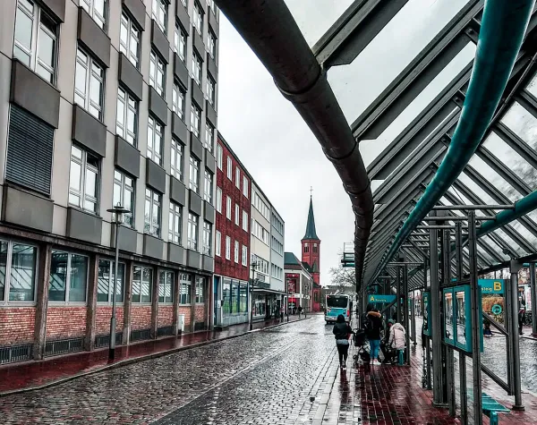 Straßenszene in Neumünster mit einem Bus, Gehweg, modernen Gebäuden und einer Kirche im Hintergrund.