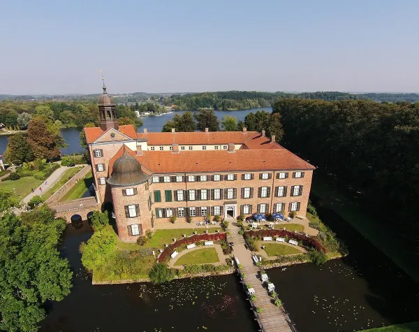 Schloss in Ostholstein, umgeben von Wasser, mit gepflegtem Garten und Brücke im Vordergrund, bei klarem Himmel.