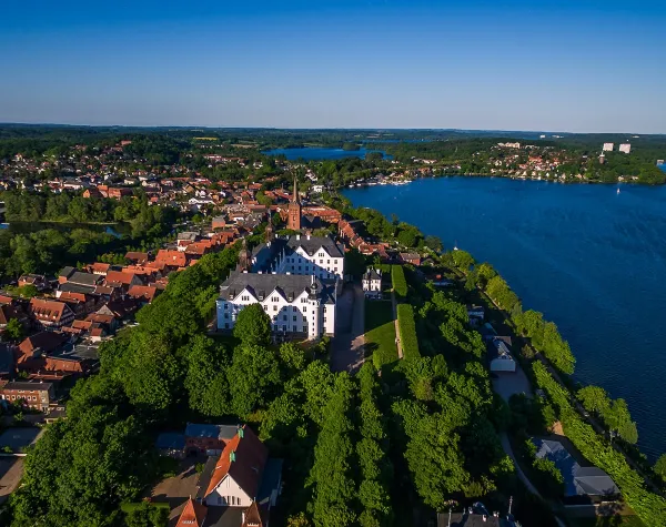 Luftaufnahme von Plön mit Plöner See im Hintergrund, umgeben von grüner Landschaft und historischen Gebäuden.